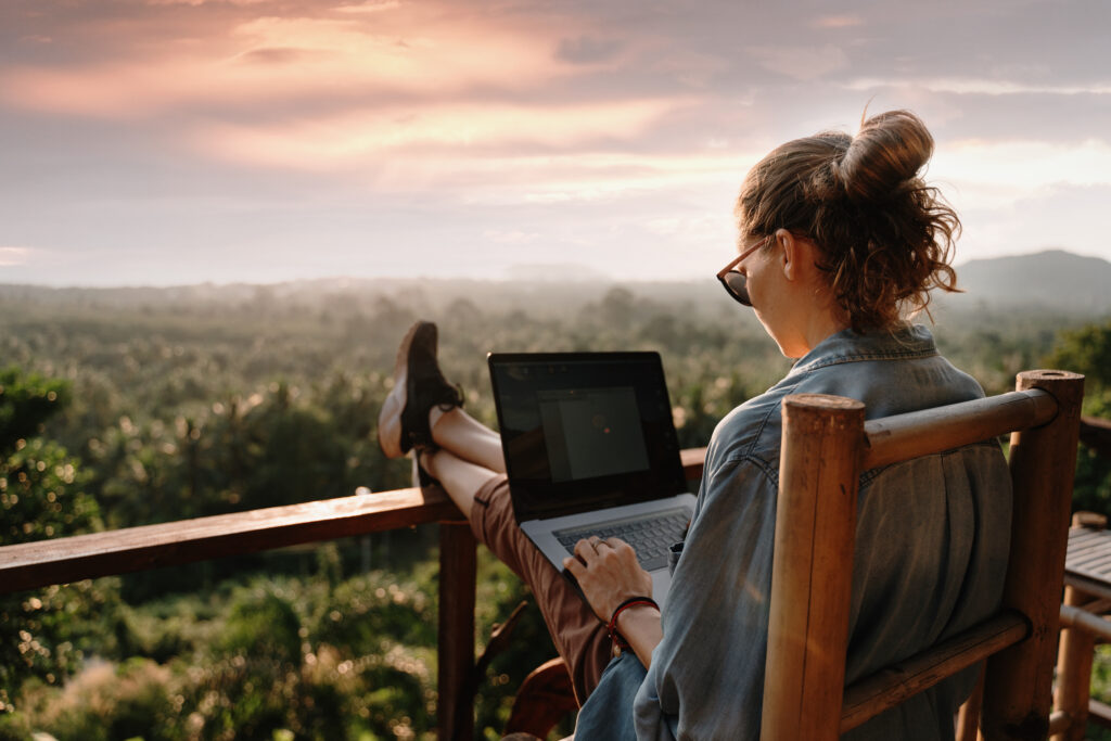A young woman working on her laptop looking out over a mountain from a deck
