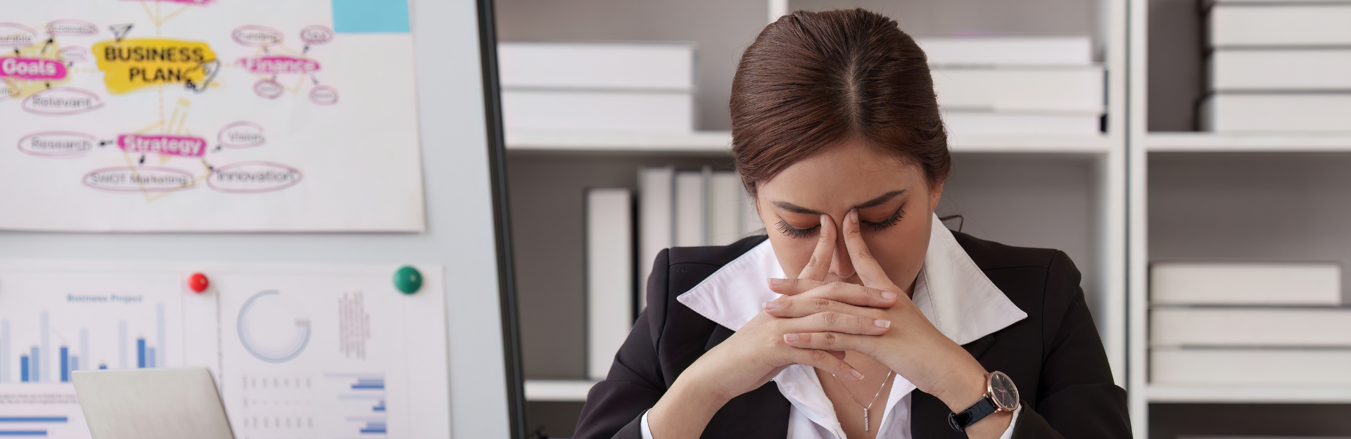 Latin woman in a business suit sitting down with hands on face looking stressed.